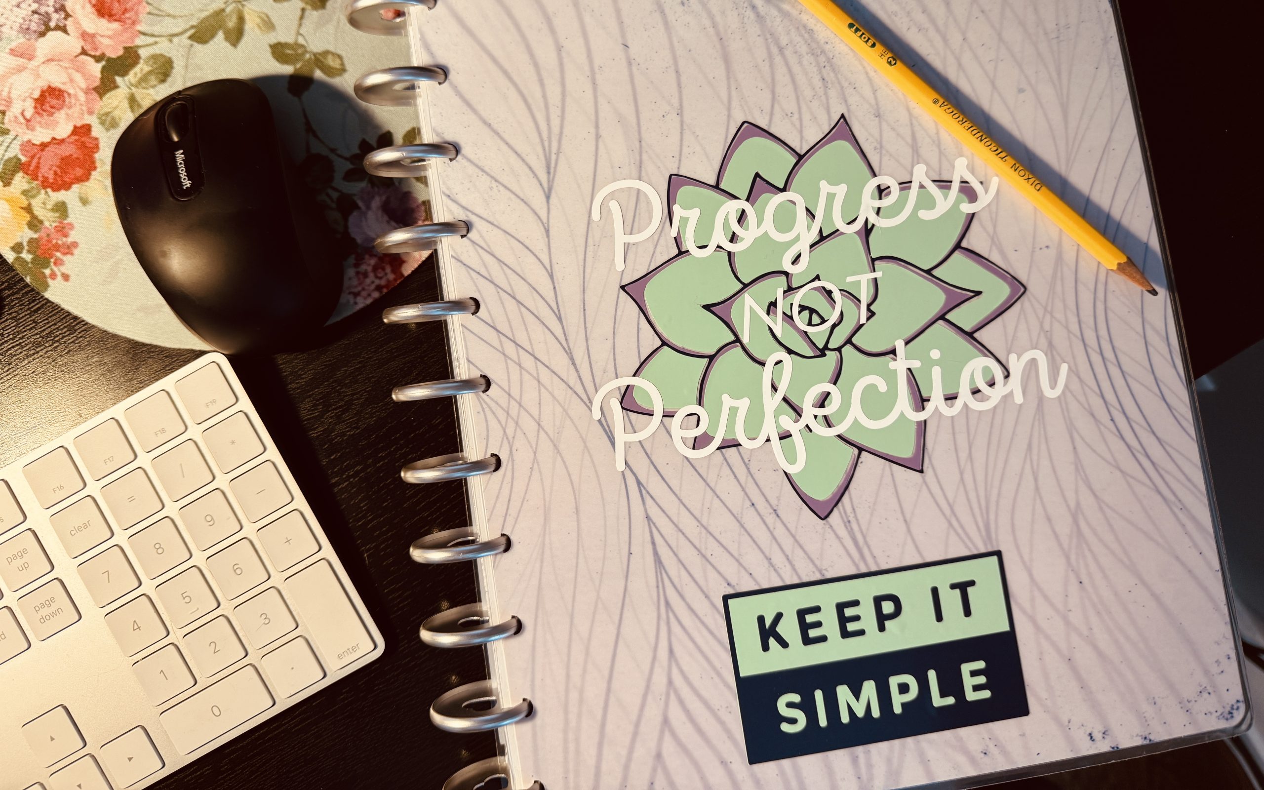 Photograph of a journal cover with the words "progress not perfection" superimposed over a decal of a succulent. At the bottom of the cover, text reads "keep it simple." There is a pencil laid at an angle across the cover. Next to the journal a portion of a computer keyboard and mouse are visible.